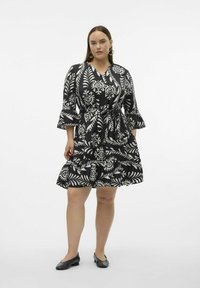 Vestido de estampado floral en blanco y negro con cintura ajustada, dobladillo con volantes y mangas acampanadas. Usado con zapatos planos negros y pendientes de aro.