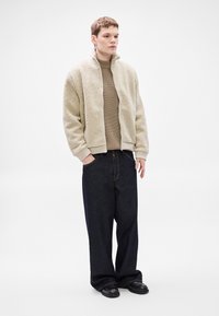 Bež flis jakna s visokim ovratnikom, rebrastim manšetama i patentnim zatvaračem sprijeda, u kombinaciji s tamnim trapericama i crnim cipelama.