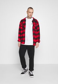 Camisa de franela a cuadros rojos y negros sobre una camiseta blanca, combinada con pantalones negros y zapatillas en blanco y negro. Diseño sencillo y ajuste casual.