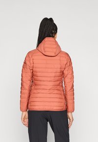 Casaco de coral almofadado com um design justo, quilting horizontal, capuz e mangas longas. Visto de trás contra um fundo claro.