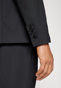 Black suit jacket sleeve featuring five black buttons. The fabric is smooth with a slight texture. Close-up of hand resting against the fabric.