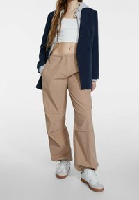 Pantaloni cargo beige con tasche laterali, indossati sotto un blazer blu navy e un hoodie grigio, abbinati a sneaker bianche con suole marroni.