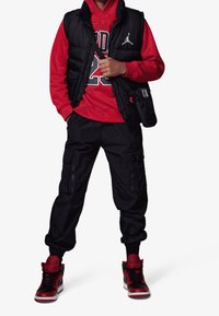 Gilet imbottito nero sopra un felpa rossa con "23" e logo Jordan, abbinato a pantaloni jogger neri e sneakers rosso e nere.