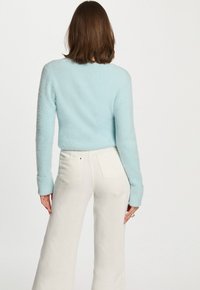 Femme portant un pull en peluche bleu clair et un pantalon large blanc taille haute, tournée vers l'arrière devant un fond blanc uni.