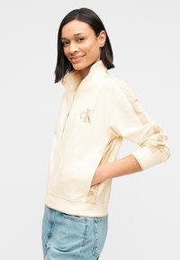 Μπεζ φούτερ με φερμουάρ και ψηλό γιακά, με χρυσό λογότυπο «CK» στο στήθος και τσέπες στα πλάγια. Λεπτή υφή, άνετη εφαρμογή.