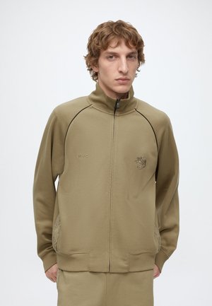 Junger Mann mit hellbraunen, lockigen Haaren trägt eine olivgrüne Jacke mit Reißverschluss und hohem Kragen sowie bestickte Logos auf der Brust.