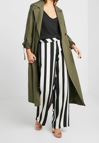 Trench-coat vert olive porté sur un haut noir, associé à un pantalon large à rayures verticales noires et blanches, et des sandales à talons transparents.