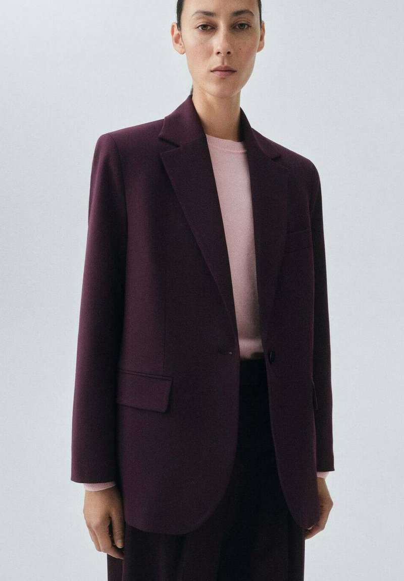 Blazer bordeaux au design ajusté avec revers crantés, fermeture à un bouton et poches latérales. Porté sur une chemise rose clair.