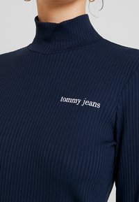Granatowy prążkowany golf z długimi rękawami, z białym haftem "tommy jeans" na lewej stronie klatki piersiowej. Gładka tekstura.