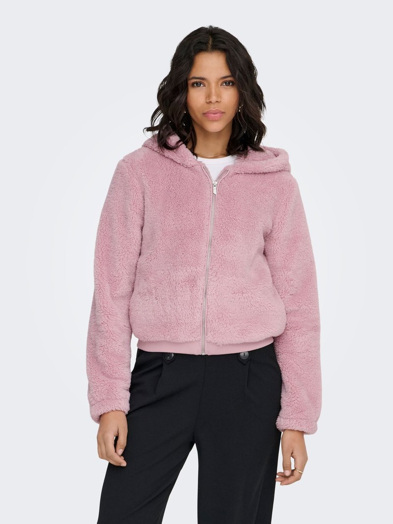 Hoodie rose à capuche en tissu duveteux avec fermeture éclair. Présente une texture douce et des poignets côtelés. Porté sur un haut blanc et associé à un pantalon noir.
