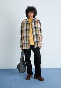 Camisa de cuadros en beige, verde y burdeos; camiseta amarilla debajo; pantalones negros; bandolera a cuadros; zapatos marrones.