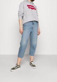 Persona che indossa un hoodie grigio Levi's, jeans corti azzurri con orlo grezzo e sneakers Vans verde oliva, in piedi su uno sfondo neutro.