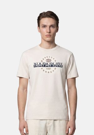 Jeune homme aux cheveux courts bruns, portant un t-shirt Napapijri beige clair avec le logo imprimé, debout devant un fond clair uni.