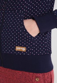 Haut à capuche zippé bleu marine avec des pois roses, présentant une poche avant et une étiquette beige. Tissu doux, poignets et ourlet inférieur côtelés.