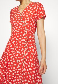 Vestido rojo de flores con mangas cortas, escote en V y botones en la parte delantera. Presenta pequeñas flores blancas sobre una tela texturizada.