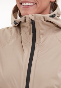 Veste imperméable beige clair avec un col haut, une fermeture éclair noire, une capuche ajustable et une texture lisse, présentant des détails de design minimalistes.
