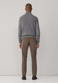 Maglione grigio con colletto alto, orlo a coste e toppe sui gomiti, abbinato a pantaloni marroni e scarpe scure, visto da dietro.