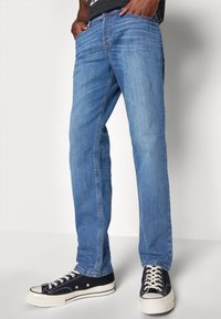 Blaue Denim-Jeans mit geradem Schnitt, die fünf Taschen und dezente Verfärbungen aufweisen. Kombiniert mit schwarzen Canvas-Sneakern.