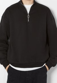 Black zip-up sweatshirt with a high neck and front zipper, made of soft fabric. Ribbed cuffs and hem; layered over a white shirt.