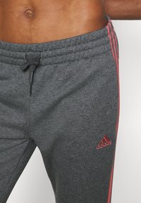 Torso masculino con pantalones deportivos Adidas gris oscuro con rayas y logotipo rojos, cordón atado en la cintura, recortado justo debajo del pecho.