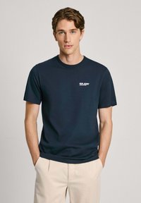 Tamsiai mėlynas medvilninis marškinėlis su apvaliu kaklu ir trumpomis rankovėmis; mažas baltas „Pepe Jeans“ logotipas kairėje krūtinės pusėje. Lygus paviršius.