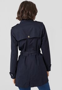 Námořnicky modrý trenčkot vyrobený z lehkého materiálu, s páskem v oblasti pasu, ramenními epauletami a větraným zadním dílem.