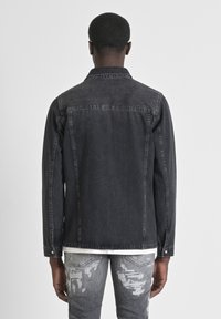 Chaqueta de mezclilla negra con cuello puntiagudo, detalles de botones y acentos de costura. Llevada sobre una camisa blanca, combinada con jeans grises desgastados.