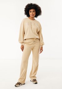 Béžová mikina se sezónními nápisy "MEXX" ozdobenými flitry, spárovaná s odpovídajícími zúženými kalhotami. Model má na sobě béžové a černé tenisky.