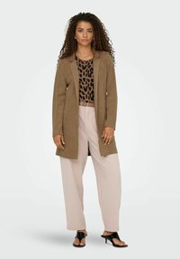 Blazer largo de ante marrón sobre una blusa estampada en negro y marrón, combinado con pantalones de pierna ancha rosa claro y sandalias de tacón negras.