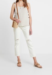 Hellbeige Leinen-Top mit Knopfdetails, kombiniert mit ausgebleichten weißen, verkürzten Jeans und braunen flachen Sandalen. Kleine gewebte Umhängetasche.
