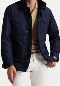 Marinblå jacka med dragkedja framtill, knäppbar krage och flera fickor framtill. Har vattenavvisande material och justerbara ärmslut.