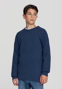 Granatowy, dzianinowy sweter o fakturze wafelka, z długimi rękawami i okrągłym dekoltem. Luźny krój, zestawiony z jasnoniebieskimi dżinsami.
