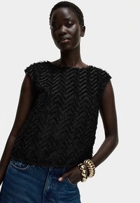 Sort tekstureret top med chevronmønster, korte ærmer og en afslappet pasform, kombineret med denim og markante gyldne armbånd.