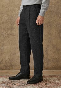 Dunkelgraue Hose mit doppelt gefaltetem Schnitt und gesprenkeltem Muster, aus strukturiertem Stoff, kombiniert mit schwarzen Lederschuhen auf einem gemusterten Teppich.