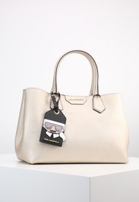 Beige syntetisk handväska med två ovandelshandtag, vinkelaktig design och en avtagbar charm med en figur i solglasögon. Guld accenter.