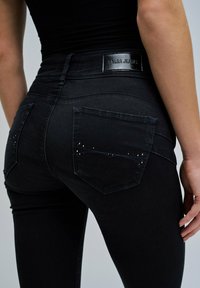 Žena nosi crne uske traperice visoki struk s metalnim detaljima na zadnjim džepovima i crnom oznakom na pojasu koja glasi "SALSA JEANS."