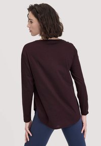 Langärmliges maroonfarbiges Oberteil aus weichem Stoff mit rundem Halsausschnitt und abgerundetem Saum. Modell kombiniert mit navy Leggings.
