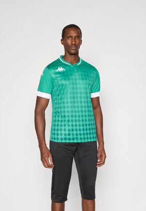 Uomo che indossa una maglietta sportiva verde a maniche corte con motivo a diamante e rifiniture bianche, pantaloni da ginnastica neri fino al ginocchio, in piedi su sfondo bianco.