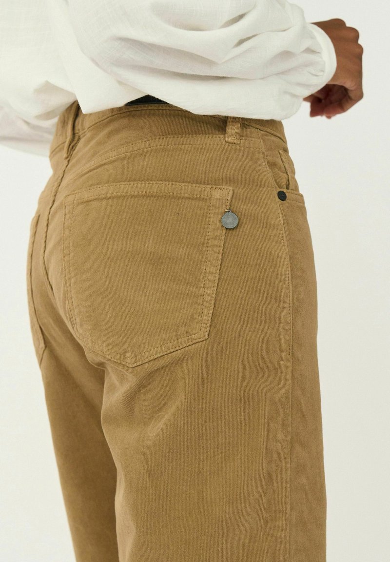 Pantalones de pana en un color marrón claro con bolsillos traseros, detalles de costura y un pequeño acento metálico cerca de la cintura.