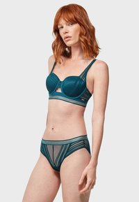 Set di reggiseno e slip in pizzo teal con coppe preformate, pannelli in rete e dettagli a righe. Il tessuto ha una finitura liscia e testurizzata con bordi ondulati.