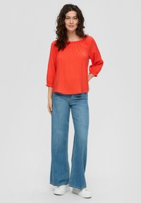 Blouse rouge texturée à manches 3/4, associée à un jean bleu à jambes larges et des baskets blanches. La blouse présente un col froncé.