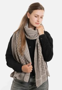 Gebreide sjaal in licht taupe met geometrische patronen, textuurstof en een franjenrand, gedragen over een zwarte trui.