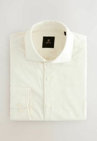 Chemise blanche pliée avec un col boutonné, présentant un tissu texturé et une étiquette noire avec "NEXT" imprimé en or.