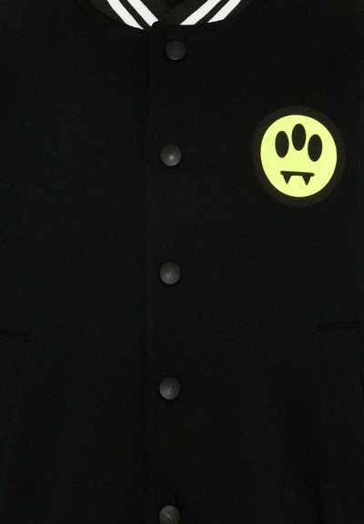 Giacca nera con bottoni, colletto a righe bianche e toppa giallo neon con un volto a tre occhi con zanne da vampiro sul petto.