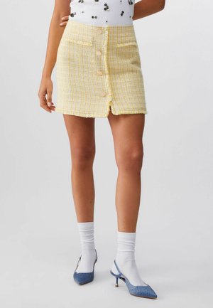 Frau trägt einen gelben Tweed-Minirock mit vorderen Knöpfen, weiße Socken und blaue Slingback-Pumps mit spitzer Spitze vor einem einfarbigen Hintergrund.