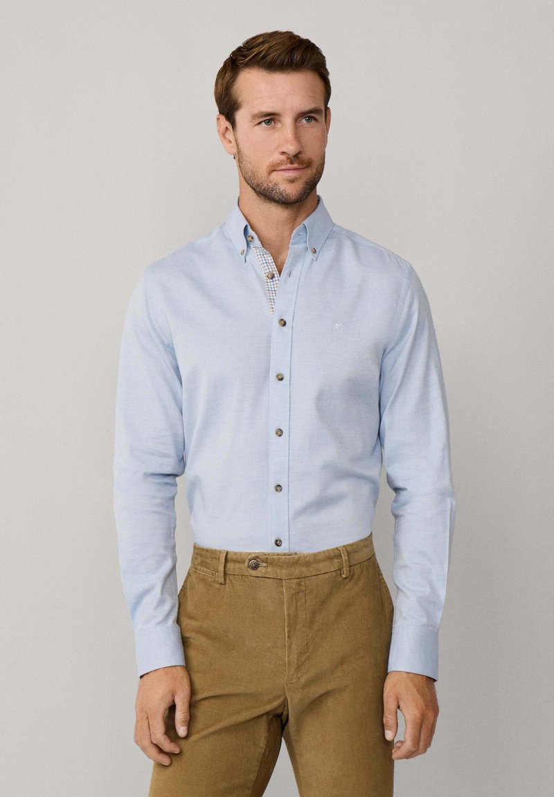 Camisa de botón azul claro con cuello, con botones oscuros y un detalle a cuadros en el interior del cuello. Combinada con pantalones de pana marrones.