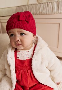 Bonnet rouge en tricot avec détail de nœud, manteau en fausse fourrure blanche, et salopette en velours côtelé rouge avec un haut à motifs comprenant des nœuds et des points.
