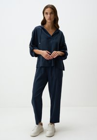 Top satin blu navy con cappuccio e maniche arrotolabili, abbinato a pantaloni coordinati e sneakers bianche, che mettono in evidenza un fit rilassato.