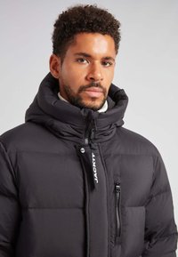 Czarna kurtka puchowa z wysokim kołnierzem, zamkiem błyskawicznym z przodu i dwoma kieszeniami po bokach. Posiada wyściełane sekcje oraz logo na uchwycie zamka.