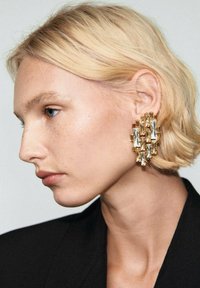 Kolczyki o złotym odcieniu, w geometrycznym kształcie, ozdobione kryształowymi akcentami i o wyraźnej strukturze. Kolczyki są duże i fakturowane, prezentujące mieszankę kształtów.